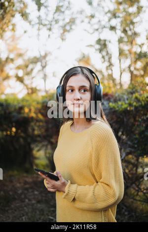 Teenager-Mädchen in gelbem Pullover mit Kopfhörern, um Musik auf ihrem Smartphone im Freien zu spielen. Stockfoto