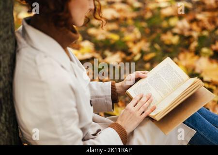 Seriöse, konzentrierte kaukasische, tausendjährige rothaarige Frau im Regenmantel liest Buch, sitzt mit gelben Blättern auf dem Boden Stockfoto