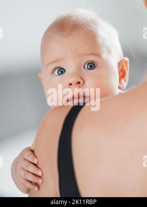 Porträt eines süßen Babys, das in den Armen der Mutter ruht, die Kamera anschaut und die Mama-Schulter berührt. Neue Mutter hält kleines Kind, umarmt Kind mit Zärtlichkeit Stockfoto