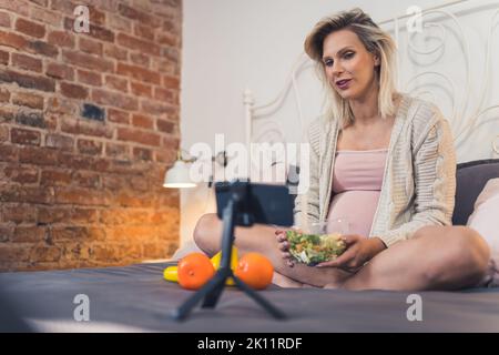 Die moderne kaukasische Schwangere sitzt auf einem Bett, isst einen nahrhaften Salat und knabert an Früchten, spricht mit ihren Freunden in einem Videoanruf. Hochwertige Fotos Stockfoto