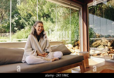 Fröhliche ältere Frau, die fröhlich lacht, während sie auf einer Couch in Lotusposition sitzt. Glückliche reife Frau, die zu Hause Meditation praktiziert. Frau genießt ein Stockfoto