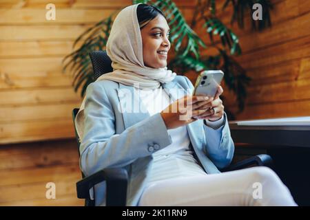 Fröhliche muslimische Geschäftsfrau lächelt, während sie eine SMS-Nachricht auf ihrem Smartphone sendet. Glückliche junge Geschäftsfrau, die an einem modernen Arbeitsplatz einen Hijab trägt. Stockfoto