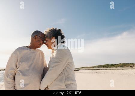 Liebevolles Seniorenpaar lächelt und berührt am Strand ihre Köpfe zusammen. Reifes Paar, das einen romantischen Moment im Freien teilt. Fröhliche ältere Leute Stockfoto