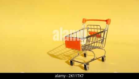 Kleiner Supermarkt Lebensmittelgeschäft Push Cart zum Einkaufen. Shopaholic. Käufer. Einkaufskonzept. Nahaufnahme. Isolierter Einkaufswagen auf gelbem Hintergrund. Cop Stockfoto