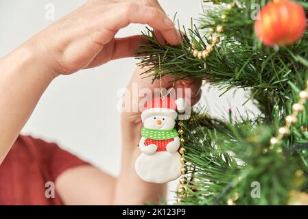 Nicht erkennbare Frau, die zu Hause einen Weihnachtsbaum schmückt, Nahaufnahme der Hände, die ein Schneemann-Ornament an einem Ast hängen. Stockfoto