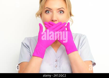 Das Porträt einer jungen kaukasischen Ärztin oder Krankenschwester ist schockiert, wenn sie ihren Mund mit ihren rosafarbenen Handschuhen vor weißem Hintergrund bedeckt Stockfoto