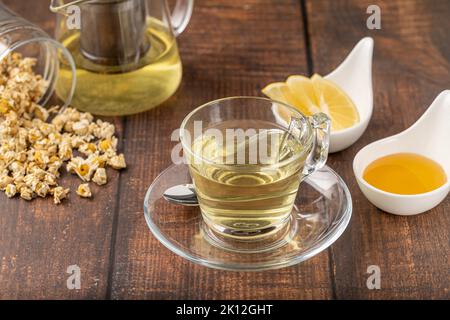 Entspannender Kamillentee in einer Glasschale mit Zitronenscheiben und Honig daneben auf einem Holztisch. Stockfoto