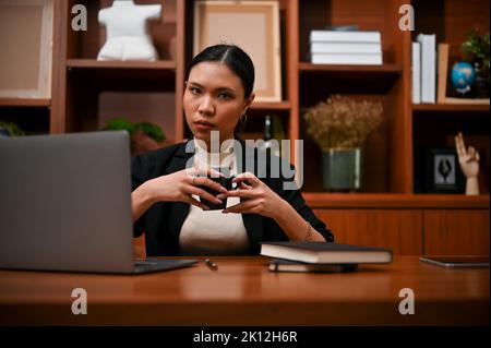 Selbstbewusste und professionelle junge asiatische Geschäftsfrau oder weibliche Chefin im formellen Anzug mit einer Kaffeetasse, die an ihrem Schreibtisch sitzt. Stockfoto