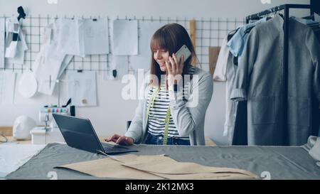Die Inhaberin eines kleinen Unternehmens bestellt Stoff für ihren Schneiderladen mit Mobiltelefon und Laptop. Frau spricht und tippt gleichzeitig. Modernes Kommunikationskonzept. Stockfoto