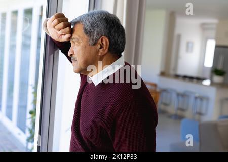 Nachdenklicher älterer Birazialmann, der sich zu Hause aus dem Wohnzimmerfenster lehnt und schaut Stockfoto