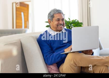 Lächelnder älterer Birazialmann mit Laptop, der im Wohnzimmer zu Hause auf der Couch sitzt Stockfoto