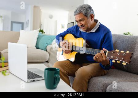 Lächelnder älterer Birazialmann, der Laptop benutzt und Gitarre spielt und auf der Couch im Wohnzimmer zu Hause sitzt Stockfoto