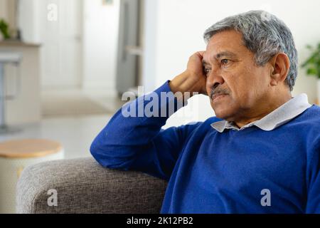 Müder, depressiver älterer Birazialmann, der auf der Couch im Wohnzimmer sitzt und sich mit dem Kopf an der Hand lehnt Stockfoto