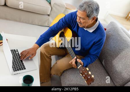 Lächelnder älterer Birazialmann, der Laptop benutzt und Gitarre spielt und auf der Couch im Wohnzimmer zu Hause sitzt Stockfoto