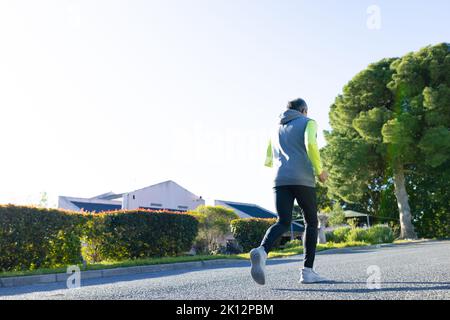 Rückansicht eines älteren Birazialmannes in Sportkleidung und Kopfhörern beim Joggen auf sonniger Straße Stockfoto