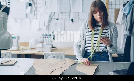 Die junge Näherin überprüft die Muster von Kleidungspapier und misst sie mit Maßband, während sie auf das Smartphone schaut. Schönes helles Studio mit Kleidungsstücken, Textilien und Nähmaschine im Hintergrund. Stockfoto