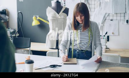 Die junge hübsche Frau zeichnet im Notizblock und schaut sich die Kleidungsskizze an, während sie in ihrem Schneideratelier arbeitet. Zeichnungen hängen an der Wand, helle Nähte sind sichtbar. Stockfoto