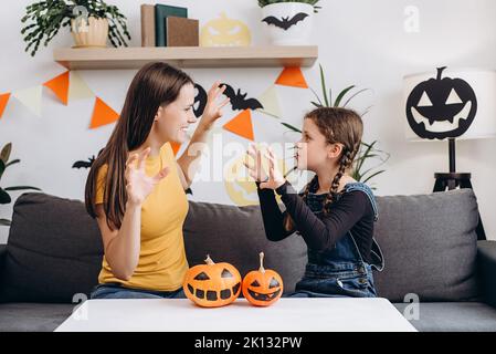 Verspielte junge Mutter, die im Wohnzimmer mit einer niedlichen kleinen Tochter auf einer bequemen Couch sitzt, erzählt gruselige Halloween-Geschichten. Lustige Mutter und Mädchen Kind haben Spaß ein Stockfoto