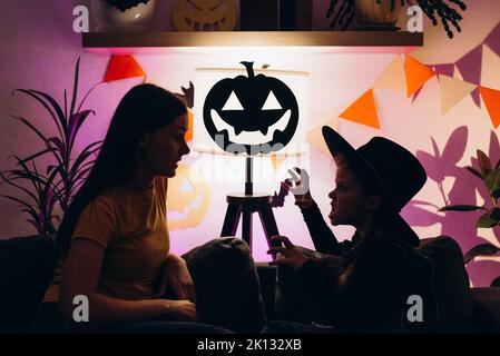 Die Silhouette einer jungen Mutter, die im Kinderzimmer mit einer niedlichen kleinen Tochter auf der Couch sitzt, erzählt gruselige Halloween-Geschichten. Lustige Mutter und Mädchen Kind haben Spaß an Stockfoto