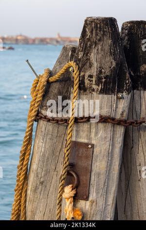 Holzsäulen mit altem Seil und Kette im Meer am Dock von Venedig. Große Holzstämme, Wellenbrecher in Venezia, Italien Stockfoto