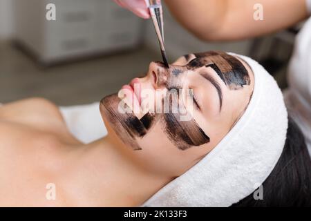 Kosmetologe, der schwarze Maske auf hübsches Frauengesicht mit schwarzen Handschuhen aufträgt, wunderschöne Frau im Spa, die Gesichtsbehandlungen hat Stockfoto