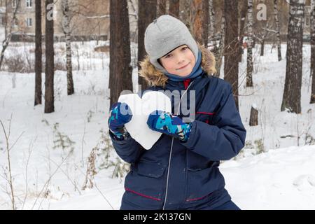 Kinder Winter. Niedlicher lächelnder Junge in einer Winterjacke und Fäustlingen, die nach einem Schneefall eine verschneite Herzform auf der Straße halten. Schnee Herz in den Händen. Stockfoto