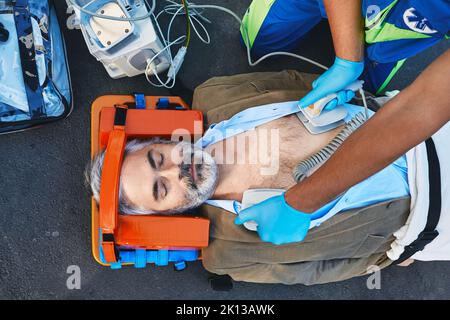 Rettungsassistent mit Defibrillator, der bei einem Unfall eine kardiopulmonale Reanimation durchführt. Erste Hilfe für verletzten reifen Mann, der in einem Notfall liegt Stockfoto