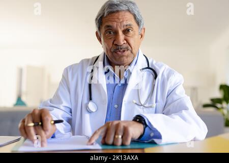 Leitender birazialer männlicher Arzt sitzt am Schreibtisch und spricht während der Videokonferenz Stockfoto