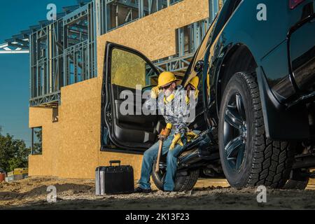 Kaukasische Auftragnehmer mittleren Alters sitzt auf Black Pickup Truck's Sill müde nach harten Tag bei der Arbeit. Modernes Wohngebäude im Bau in Stockfoto