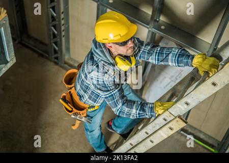 Foto von einem professionellen Auftragnehmer in gelber Hartmütze, der die Leiter auf der Baustelle besteigte. Verwendete Bauwerkzeuge und -Geräte. Stockfoto