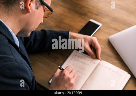 Horizontale Aufnahme von oben des Geschäftsmannes, der morgens mit Notizen aus seinem Notizbuch beschäftigt war. Mann trägt Brille füllt Notebook mit Stift, organisieren Stockfoto