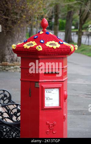 Skurriler, farbenfroher, handgestrickter Wollhut ziert in Solidarität mit der Ukraine eine alte rote viktorianische Säulenkiste mit gehäkelten Sonnenblumen. VEREINIGTES KÖNIGREICH. Stockfoto