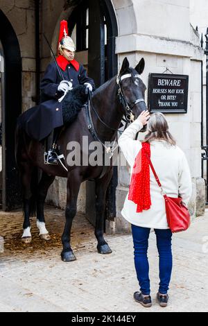 Eine Frau streichelt bei der Horseguards Parade auf das Pferd eines Soldaten, im Hintergrund ein Schild mit der Aufschrift „Pferde dürfen treten oder beißen“, Whitehall, London, Großbritannien Stockfoto