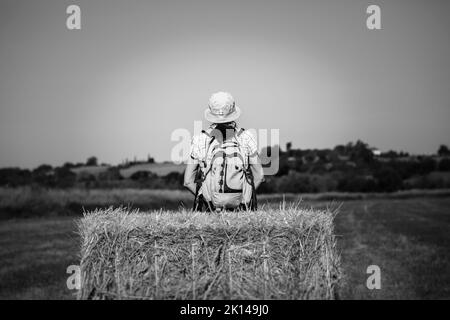 Schwarz-weißes Rückbild einer Wanderfrau mit Rucksack und Sommerhut, die isoliert auf einem Heuballen sitzt und in die offene Landschaft Großbritanniens blickt. Stockfoto