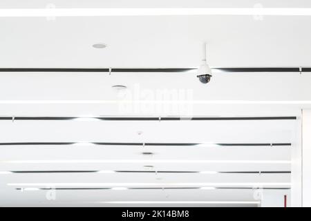 Weiße CCTV-Kamera mit Kuppel aus Acrylglas in der Mitte, die von der Decke hängt. Der Hintergrund ist weißes Studio. Stockfoto