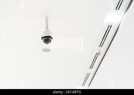 Weiße CCTV-Kamera mit Kuppel aus Acrylglas in der Mitte, die von der Decke hängt. Der Hintergrund ist weißes Studio. Stockfoto