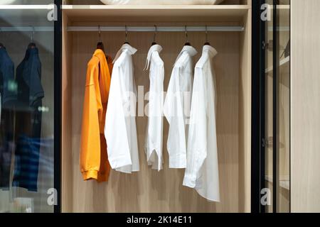 Hemden hängen in einem begehbaren Schrank mit Glastür. Eine davon ist orange und unterscheidet sich von anderen, die weiß sind. Stockfoto