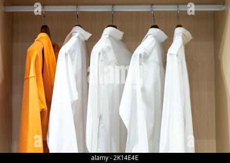 Hemden hängen in einem begehbaren Schrank mit Glastür. Eine davon ist orange und unterscheidet sich von anderen, die weiß sind. Stockfoto
