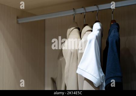 Hemden hängen in einem begehbaren Schrank mit Glastür. Eine davon ist in Weiß gehalten, die sich von anderen in Beige und Dunkelblau absetzt. Stockfoto