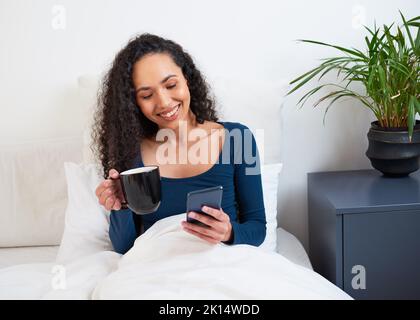 Eine junge, multiethnische Frau trinkt Kaffee im Bett, während sie am Telefon aufwacht Stockfoto