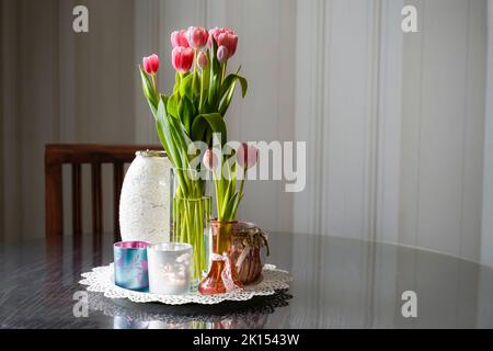 Langstielige rosa Tulpen mit mehreren Gläsern auf einem runden Holztisch. Im Hintergrund befindet sich ein Stuhl und eine gestreifte Tapete. Antik-romantischer Look. Stockfoto