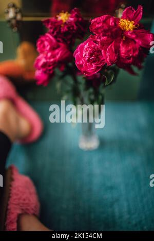 Sterbende magenta-rosa Pfingstrosen-Blüten in Kristallvase auf blauem Teppich mit Frau in rosa flauschigen Hausschuhen Stockfoto