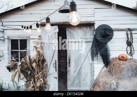 Glühbirnen auf der Girlande in der Nähe einer alten, gruselig geschmückten Scheune mit Heu, Heuhaufen. Feier von halloween. Kürbis-Jack-o-Laterne. Hexenkostüm, Hut.Hor Stockfoto