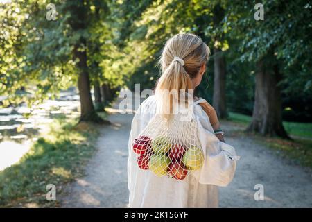Frau, die im öffentlichen Park einen Netzbeutel mit Äpfeln hält. Nachhaltiger Lebensstil und Stadtleben Stockfoto
