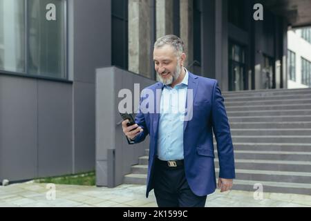 Älterer grauhaariger, hübscher Mann in einem Anzug. Er geht in die Nähe des Bürogebäudes, hält das Telefon in den Händen, macht einen Videoanruf, spricht, lächelt. Stockfoto