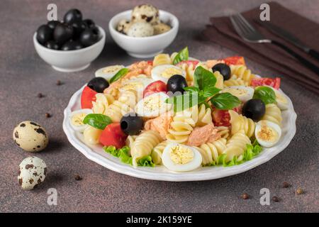 Lachssalat aus der Dose mit Pasta, Kirschtomaten, Wachteleiern, Basilikum und schwarzen Oliven auf braunem Hintergrund, Nahaufnahme Stockfoto
