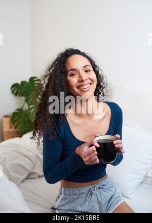 Eine glückliche junge, multiethnische Frau wacht auf, indem sie im Schlafzimmer Kaffee trinkt Stockfoto