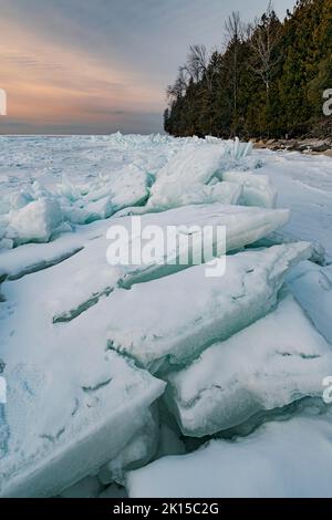 Das Licht des Sonnenuntergangs färbt den Himmel über Eismustern und Haufen von Eisplatten, die Eisschufen genannt werden, versammeln sich am Ufer des Lake Michigan in den Gewässern der Green Bay Stockfoto