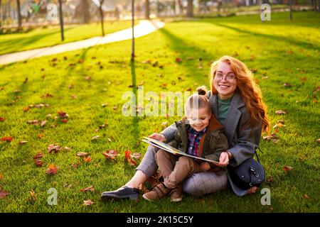 Rotschopf Frau liest Buch mit kleinen Jungen auf Gras im Stadtpark sitzen. Die junge Mutter unterrichtet ihren Sohn mit der ersten Klasse und macht Hausaufgaben am sonnigen Herbsttag Stockfoto