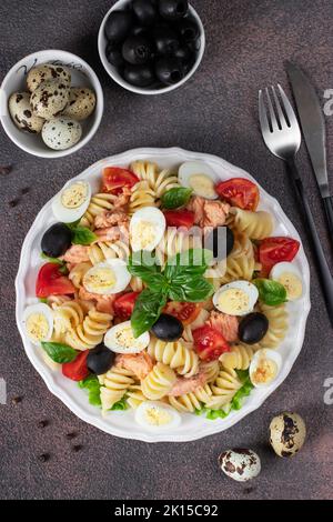 Lachssalat aus der Dose mit Pasta, Kirschtomaten, Wachteleiern, Basilikum und schwarzen Oliven auf braunem Hintergrund Stockfoto